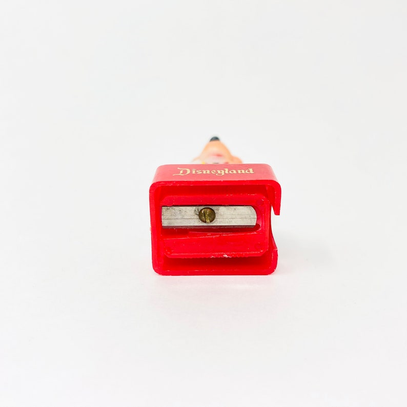 May include: A red pencil sharpener with the word "Disneyland" in gold lettering. The sharpener has a silver blade and a pencil tip extending from the top. The background is white.