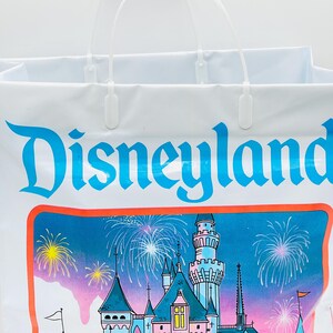 May include: A white plastic shopping bag featuring the blue Disneyland logo at the top. Below the text is a colorful illustration of a castle with pink and blue towers, surrounded by bright yellow and white fireworks against a blue sky.