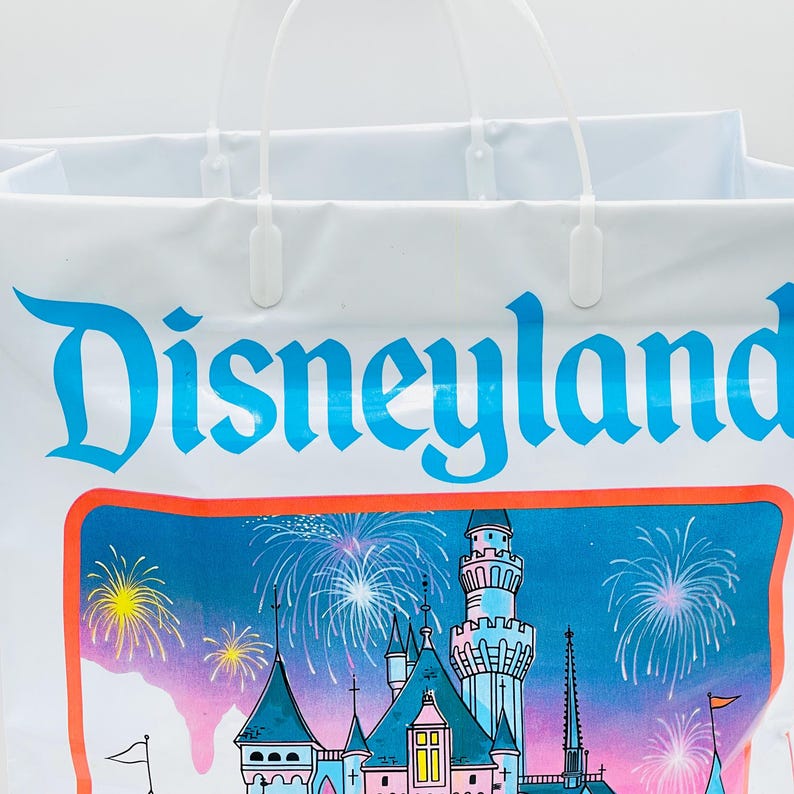 May include: A white plastic shopping bag featuring the blue Disneyland logo at the top. Below the text is a colorful illustration of a castle with pink and blue towers, surrounded by bright yellow and white fireworks against a blue sky.