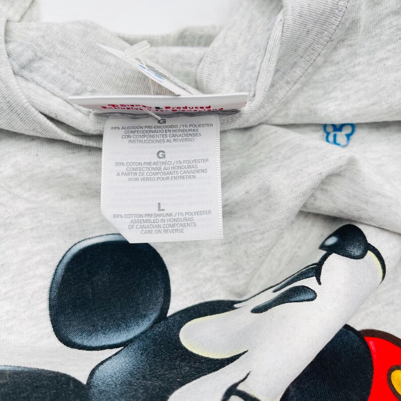 May include: A light gray heathered t-shirt featuring a large graphic of Mickey Mouse. A white fabric care label is attached to the inner neckline, displaying text in English, French, and Spanish, including size L and material composition details.