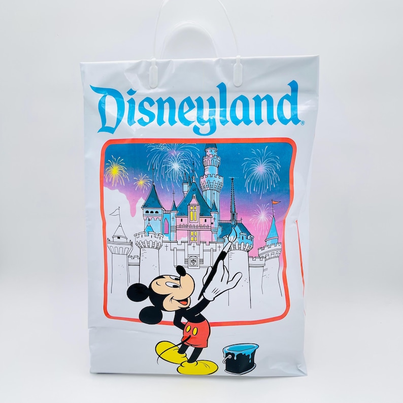 May include: A white plastic shopping bag featuring the blue Disneyland logo at the top. The center graphic shows Mickey Mouse painting a castle with fireworks in the background, with a small blue paint bucket sitting on the ground nearby.
