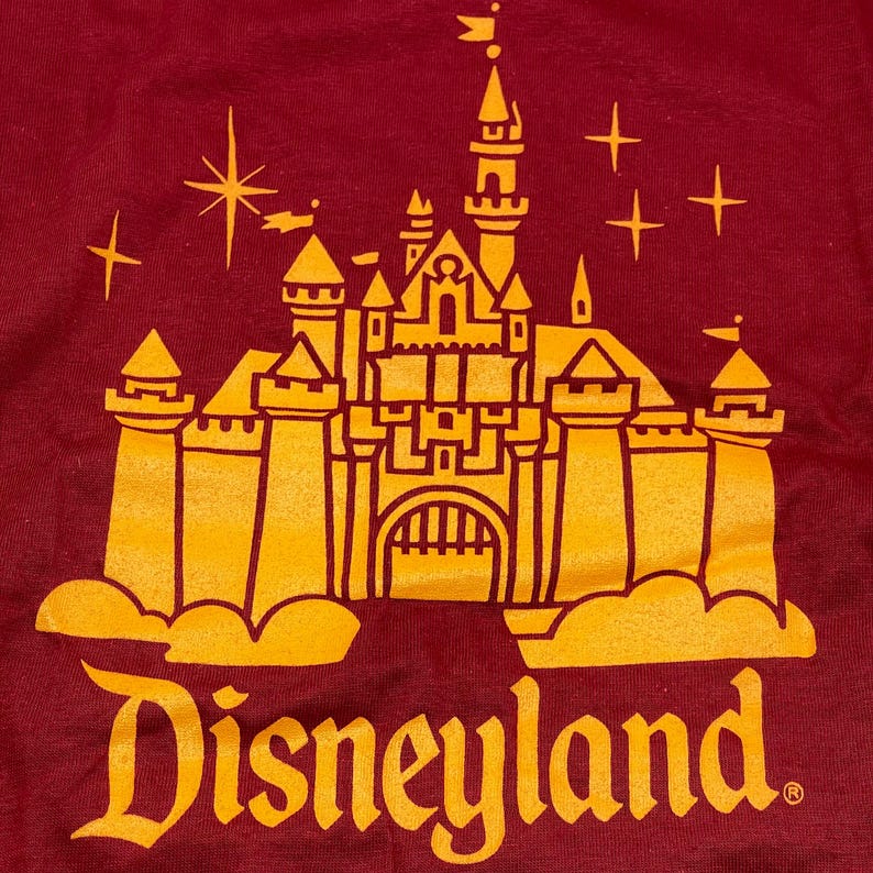 May include: A dark red fabric featuring a screen-printed graphic in yellow ink. The design depicts a detailed castle with multiple towers, spires, and flags, surrounded by small star shapes. The word Disneyland is printed in a bold font below.
