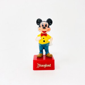 May include: A vintage Mickey Mouse figurine on a red base with gold "Disneyland" text. Mickey wears a yellow shirt, blue trousers, a red bow tie, and brown shoes. The figurine has black ears and a smiling face, a classic Disney collectible.