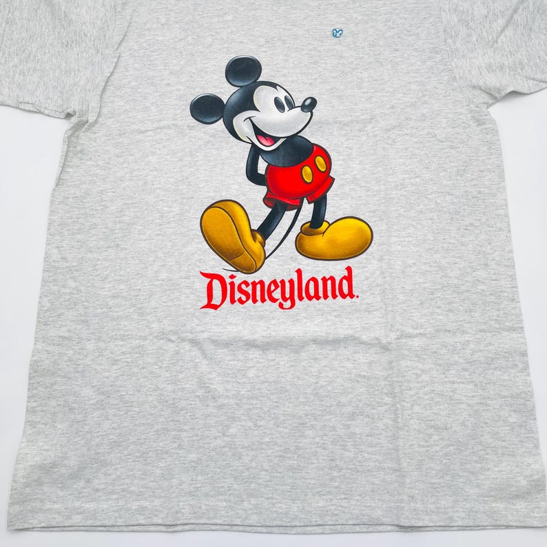 May include: A light gray heathered t-shirt featuring a graphic of Mickey Mouse wearing red shorts and yellow shoes. Below the character, the word Disneyland is printed in a bold red serif font. The design includes a small blue icon above.