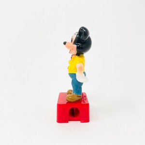 May include: A vintage Mickey Mouse pencil sharpener. The figure is wearing a yellow shirt, blue trousers, and brown shoes. The sharpener is red and has a hole for the pencil. The figure is standing on the red base.