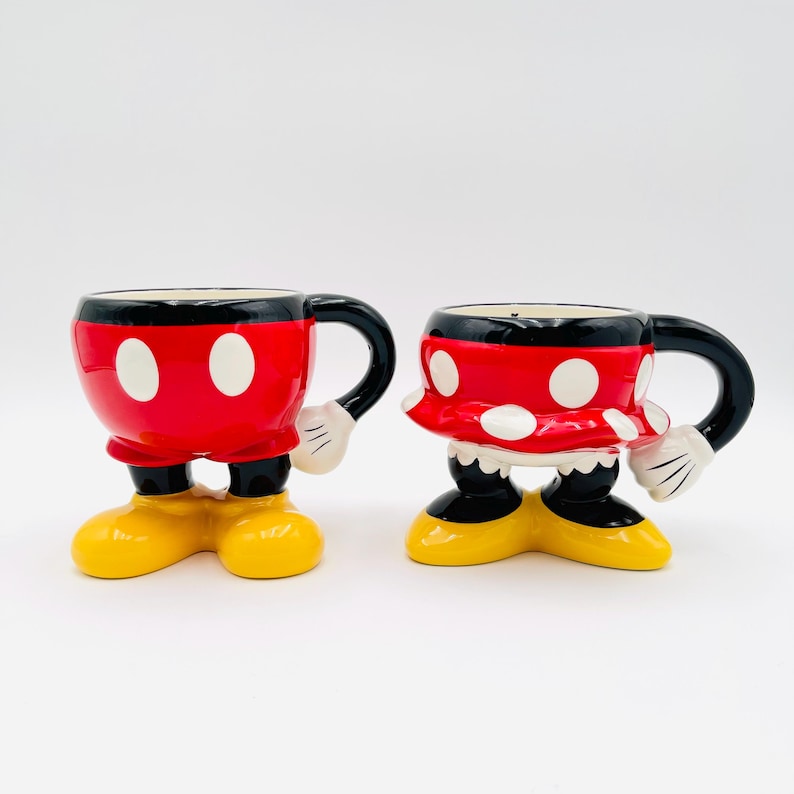 Op de afbeelding: Twee keramische mokken, &eacute;&eacute;n rood met witte stippen en de andere rood met witte stippen, gevormd als stripfiguren. Elke mok heeft zwarte handvatten en gele voeten. De mokken zijn ontworpen om op een populair stripfiguurpaar te lijken.