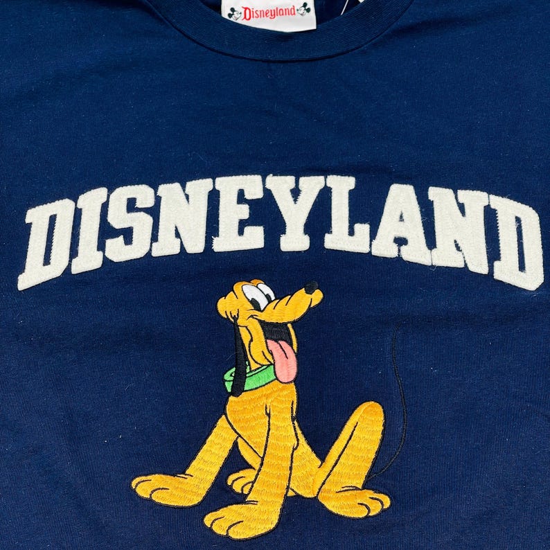 May include: A navy blue t-shirt featuring the white text Disneyland arched across the chest. Below the text is an embroidered graphic of the yellow dog Pluto with a green collar and a pink tongue, with a small Disneyland tag at the neckline.