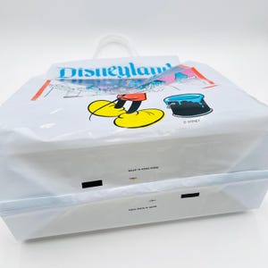 May include: A white plastic shopping bag featuring the blue Disneyland logo, a graphic of Mickey Mouse's yellow shoes, and a blue paint bucket. The bag includes the text Disney, Made in Hong Kong, and has a white plastic handle at the top.