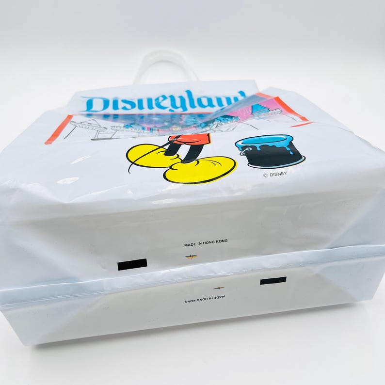 May include: A white plastic shopping bag featuring the blue Disneyland logo, a graphic of Mickey Mouse's yellow shoes, and a blue paint bucket. The bag includes the text Disney, Made in Hong Kong, and has a white plastic handle at the top.