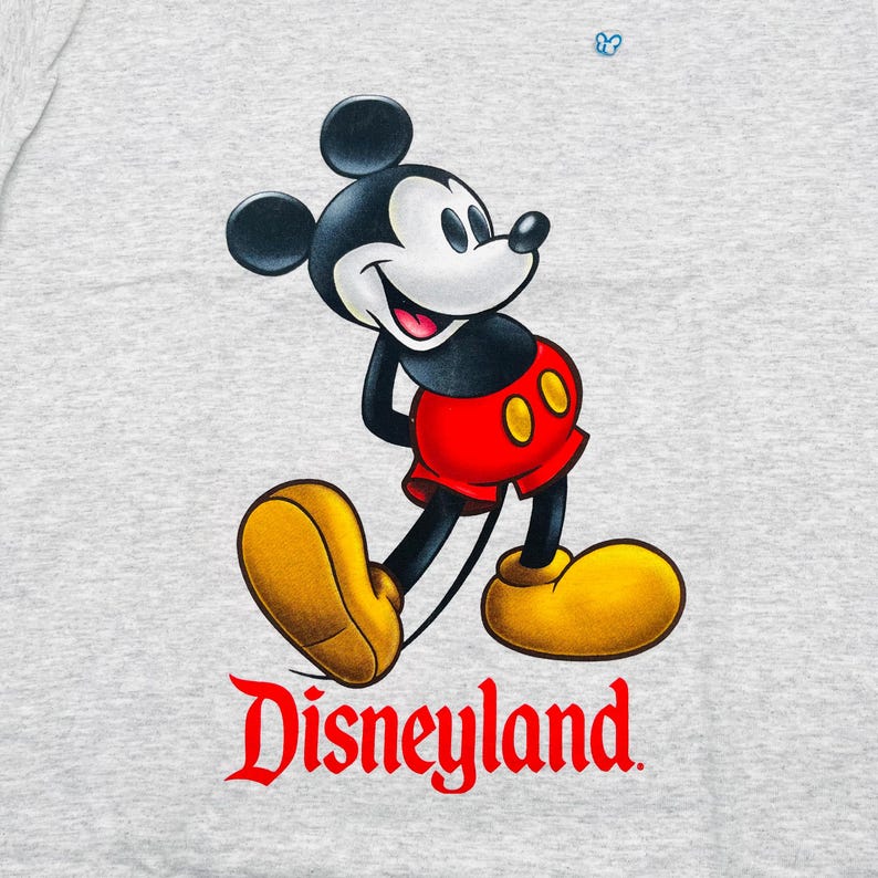 May include: A light gray heathered t-shirt featuring a graphic of Mickey Mouse wearing red shorts and yellow shoes. The character is positioned above the red text that reads Disneyland. A small blue icon is visible above the character's head.