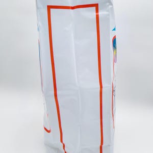 May include: A tall white rectangular plastic bag featuring a prominent vertical orange rectangular border on the front surface. The sides of the bag display partial blue and red graphic elements with thin line drawings of fireworks and star shapes.
