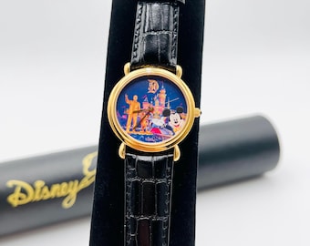 May include: Gold-tone watch with a black leather band. The watch face features a colorful image of a castle, Mickey Mouse, and other characters. The watch is a Disney 50th anniversary edition.