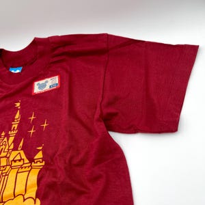 May include: A maroon short-sleeve t-shirt featuring a yellow graphic of a castle with stars. A rectangular tag is attached to the chest area, displaying a blue Mickey Mouse icon, the text ADULT LARGE, and other small printed details.