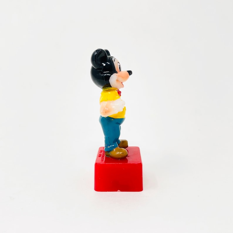 May include: A small, vintage Mickey Mouse figurine. The figure is wearing a yellow shirt, blue trousers, and brown shoes. It stands on a red rectangular base. The figurine has black ears and a red bow tie.
