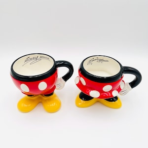 Op de afbeelding: Twee keramische mokken in de vorm van Mickey en Minnie Mouse. De Mickey-mok is rood met witte stippen, zwarte rand en gele voeten. De Minnie-mok is rood met witte stippen, zwarte rand en gele voeten. Elke mok heeft een zwart handvat en de naam van het personage.