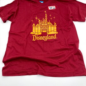 May include: A maroon short-sleeved t-shirt featuring a yellow graphic of a castle with the text Disneyland below it. The shirt includes a small rectangular tag near the neckline with a Mickey Mouse icon and the word Adult printed on it.