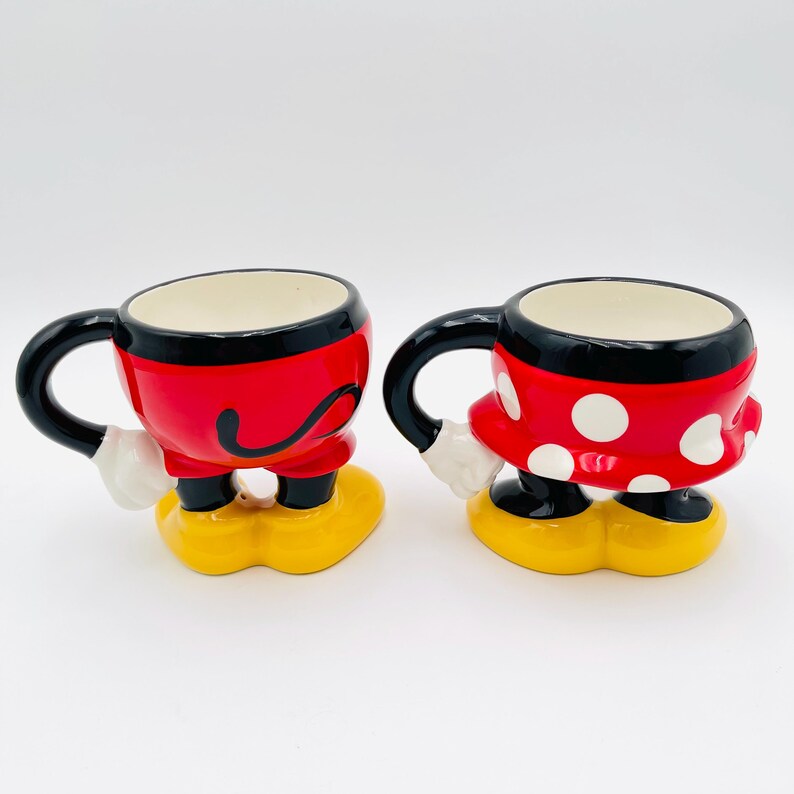 Op de afbeelding: Twee keramische mokken, &eacute;&eacute;n rood met zwarte accenten en de andere rood met witte stippen. Beide mokken hebben zwarte handvatten, witte handschoenen en gele voeten, die op stripfiguren lijken. De mokken zijn ontworpen om op Mickey en Minnie Mouse te lijken.