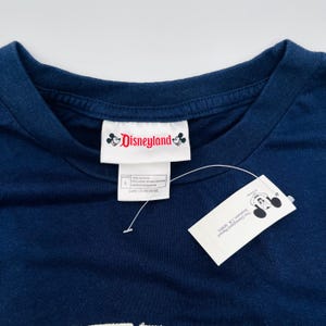 May include: A dark blue t-shirt featuring a white inner neck label with the red Disneyland logo and Mickey Mouse icons. A white paper hang tag is attached with a plastic fastener, displaying the Disneyland Resort Anaheim, CA 92803 address.