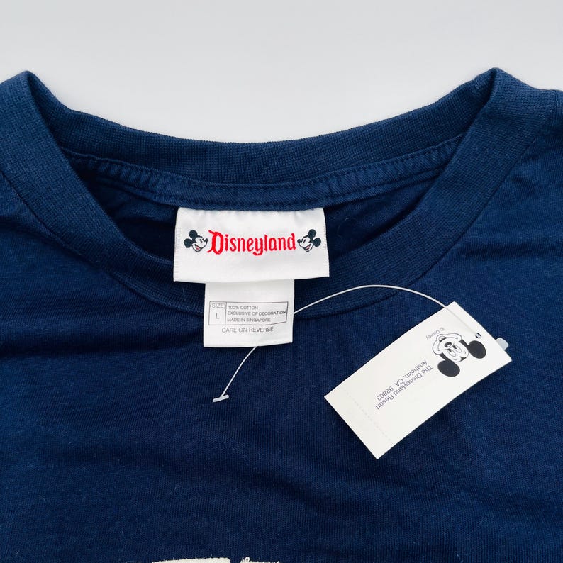 May include: A dark blue t-shirt featuring a white inner neck label with the red Disneyland logo and Mickey Mouse icons. A white paper hang tag is attached with a plastic fastener, displaying the Disneyland Resort Anaheim, CA 92803 address.
