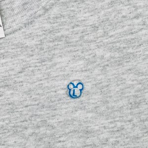 May include: A light gray heathered fabric t-shirt featuring a small, blue, circular embroidered logo. The logo depicts a stylized outline of a mouse head with the capital letter L centered inside. A white tag with the number 4 is visible at the top.