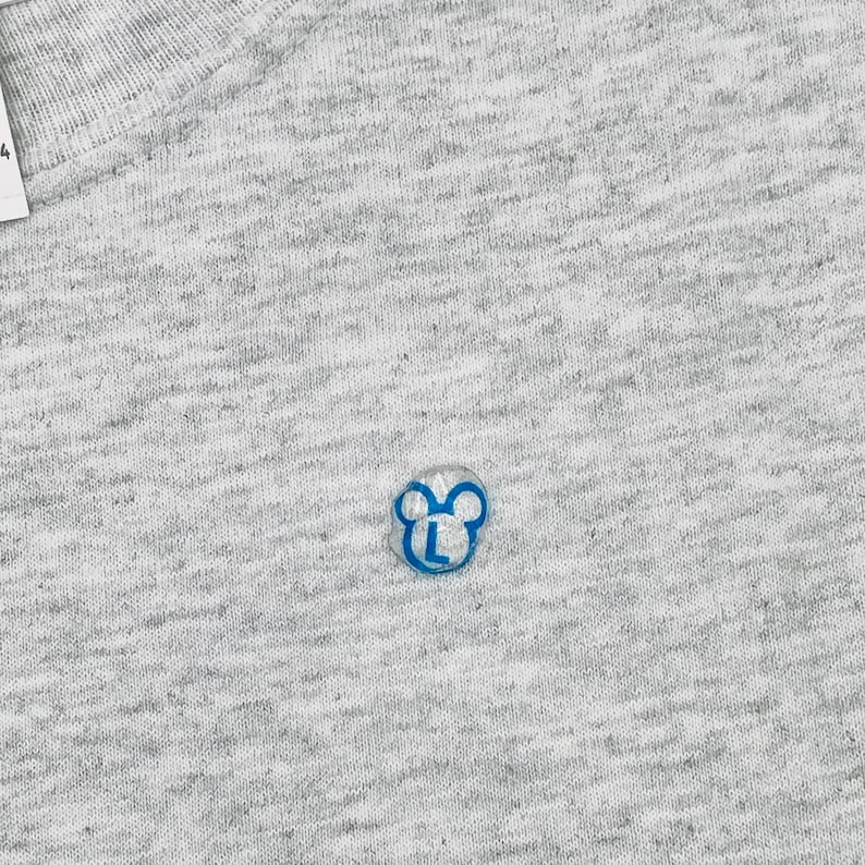 May include: A light gray heathered fabric t-shirt featuring a small, blue, circular embroidered logo. The logo depicts a stylized outline of a mouse head with the capital letter L centered inside. A white tag with the number 4 is visible at the top.