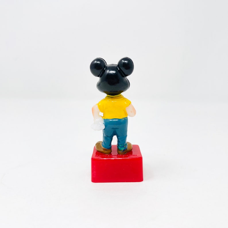May include: A small figurine of a cartoon mouse, viewed from the back. The mouse has a black head, a yellow shirt, blue trousers, and brown shoes. It stands on a red rectangular base.