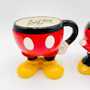 Op de afbeelding: Een keramische Mickey Mouse mok, rood met witte stippen en een zwarte rand. Het heeft een zwart handvat en gele voeten. De tekst "Mickey Mouse" staat aan de binnenkant. Een verzamelobject.