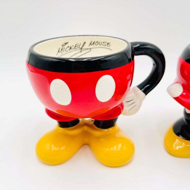 Op de afbeelding: Een keramische Mickey Mouse mok, rood met witte stippen en een zwarte rand. Het heeft een zwart handvat en gele voeten. De tekst "Mickey Mouse" staat aan de binnenkant. Een verzamelobject.