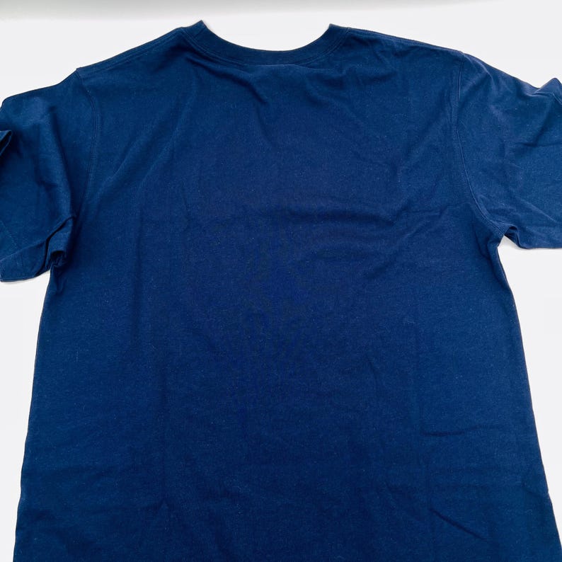 May include: A solid navy blue short-sleeved t-shirt with a crew neckline. The garment is displayed flat, showing the plain back side of the shirt. The fabric appears to be a standard cotton blend with a smooth, consistent texture throughout the piece.