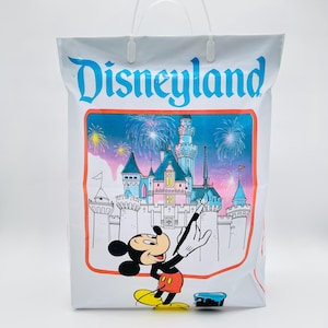 May include: A white plastic shopping bag featuring the blue Disneyland logo at the top. The center graphic shows Mickey Mouse painting a castle with fireworks in the background, rendered in shades of blue, pink, and yellow with a red border.