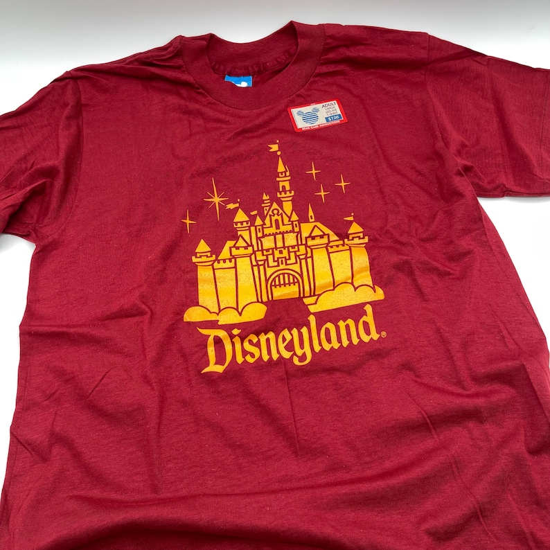 May include: A maroon short-sleeved t-shirt featuring a gold graphic of a castle with the text Disneyland below it. The shirt includes a small rectangular tag near the neckline with a Mickey Mouse icon and the text Adult.