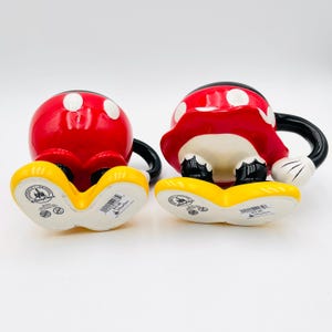 Op de afbeelding: Twee Mickey Mouse mokken van keramiek. De mokken zijn rood met witte stippen, zwarte handvatten en gele voeten. De mokken hebben de vorm van Mickey Mouses hoofd en voeten. De mokken staan op een witte achtergrond.