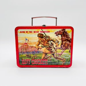 May include: Red metal lunchbox with a picture of Davy Crockett on the Alamo on the front. The text "King of the Wild Frontier" and "Davy Crockett at the Alamo" are printed on the lunchbox.