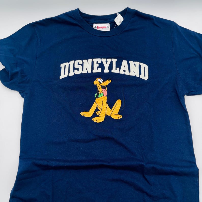 May include: A navy blue short-sleeve t-shirt featuring the word DISNEYLAND in white arched lettering across the chest. Below the text is a graphic of the yellow dog character Pluto wearing a green collar, with a white tag attached to the collar.