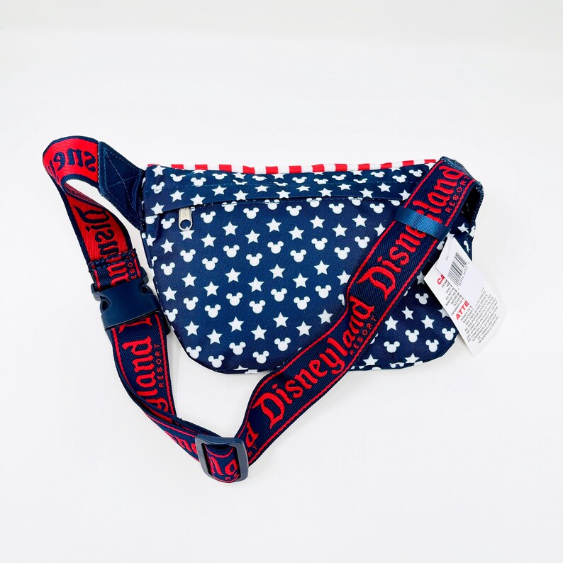 Puede incluir: Ri&ntilde;onera azul marino con un estampado de estrellas blancas y cabezas de Mickey Mouse. La correa roja lleva la inscripci&oacute;n "Disneyland Resort" en blanco. El borde superior tiene detalles en rojo. Lleva una peque&ntilde;a etiqueta.