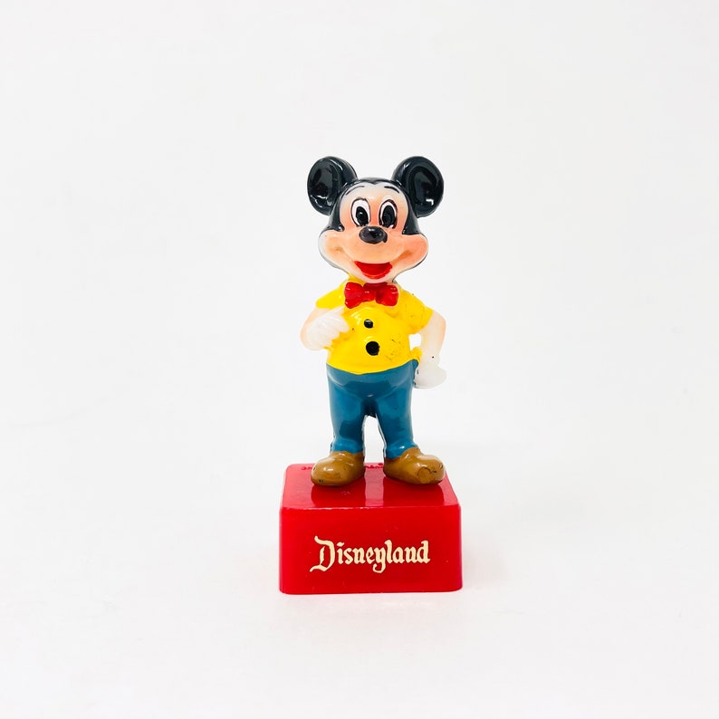 May include: A vintage Mickey Mouse figurine on a red base. Mickey wears a yellow shirt, blue trousers, a red bow tie, and brown shoes. The base reads "Disneyland" in gold lettering.