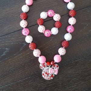 May include: A pink and red beaded necklace and bracelet set with a Minnie Mouse charm. The beads are white with red hearts, pink, and red.