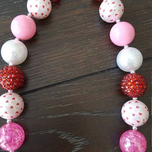 May include: A pink, white, and red chunky necklace with a silver clasp. The beads are made of different materials, including plastic, glitter, and fabric. The necklace is perfect for a Valentine's Day party or a special occasion.