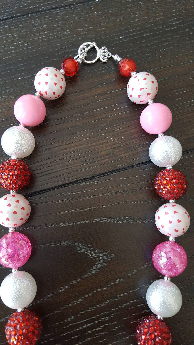May include: A pink, white, and red chunky necklace with a silver clasp. The beads are made of different materials, including plastic, glitter, and fabric. The necklace is perfect for a Valentine's Day party or a special occasion.