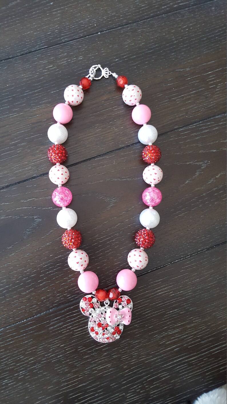 May include: A pink, white, and red beaded necklace with a rhinestone Minnie Mouse charm. The beads are various sizes and have different patterns, including hearts and polka dots.