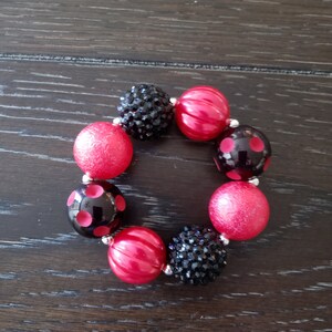 May include: A red and black beaded bracelet with a variety of textures and patterns. The bracelet features red beads with a smooth finish, red beads with a textured finish, black beads with a polka dot pattern, and black beads with a sparkly finish.