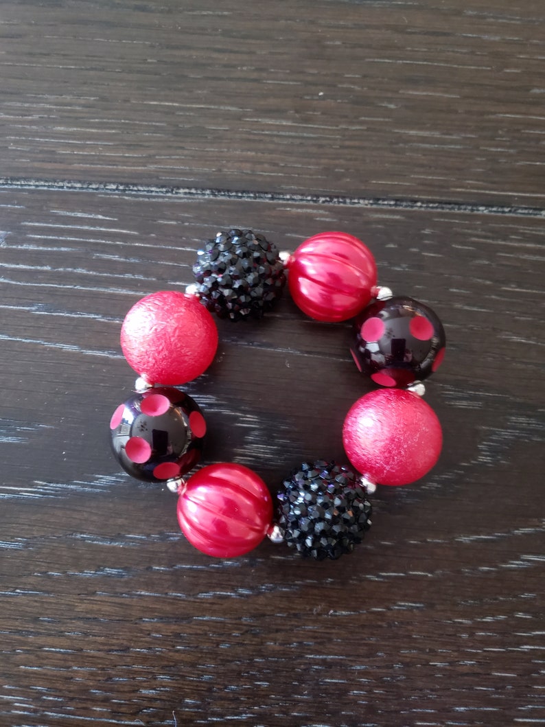 May include: A red and black beaded bracelet with a variety of textures and patterns. The bracelet features red beads with a smooth finish, red beads with a textured finish, black beads with a polka dot pattern, and black beads with a sparkly finish.