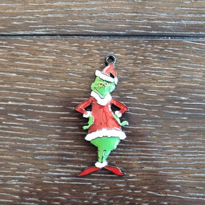 May include: A green Grinch Christmas ornament with a red suit, white fur trim, and a Santa hat. The ornament is made of metal and has a black wire loop for hanging.