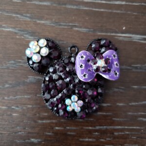 Pu&ograve; includere: Un ciondolo Mickey Mouse in strass nero e viola con un fiocco viola. Il ciondolo &egrave; in metallo e ha un anello per attaccarlo a una collana o un braccialetto.