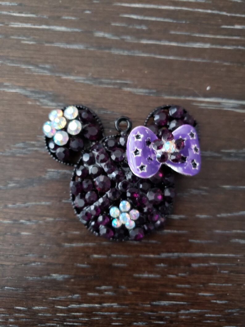 Pu&ograve; includere: Un ciondolo Mickey Mouse in strass nero e viola con un fiocco viola. Il ciondolo &egrave; in metallo e ha un anello per attaccarlo a una collana o un braccialetto.