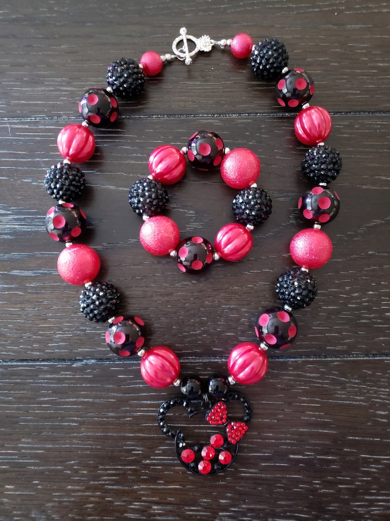 May include: A red and black beaded necklace and bracelet set with a Minnie Mouse charm. The necklace and bracelet are made with large, chunky beads and have a silver clasp.
