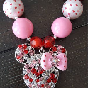 May include: A pink and red chunky bead necklace with a rhinestone Minnie Mouse charm. The necklace is made with large, round beads and features a silver chain with a pink bow.