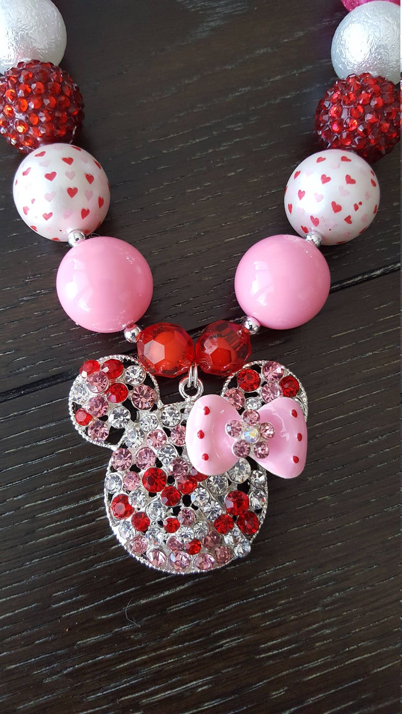 May include: A pink and red chunky bead necklace with a rhinestone Minnie Mouse charm. The necklace is made with large, round beads and features a silver chain with a pink bow.