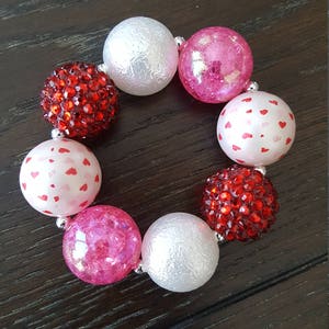 May include: A Valentine's Day themed bracelet with red and pink beads. The beads are made of different materials, including glitter, hearts, and textured plastic.