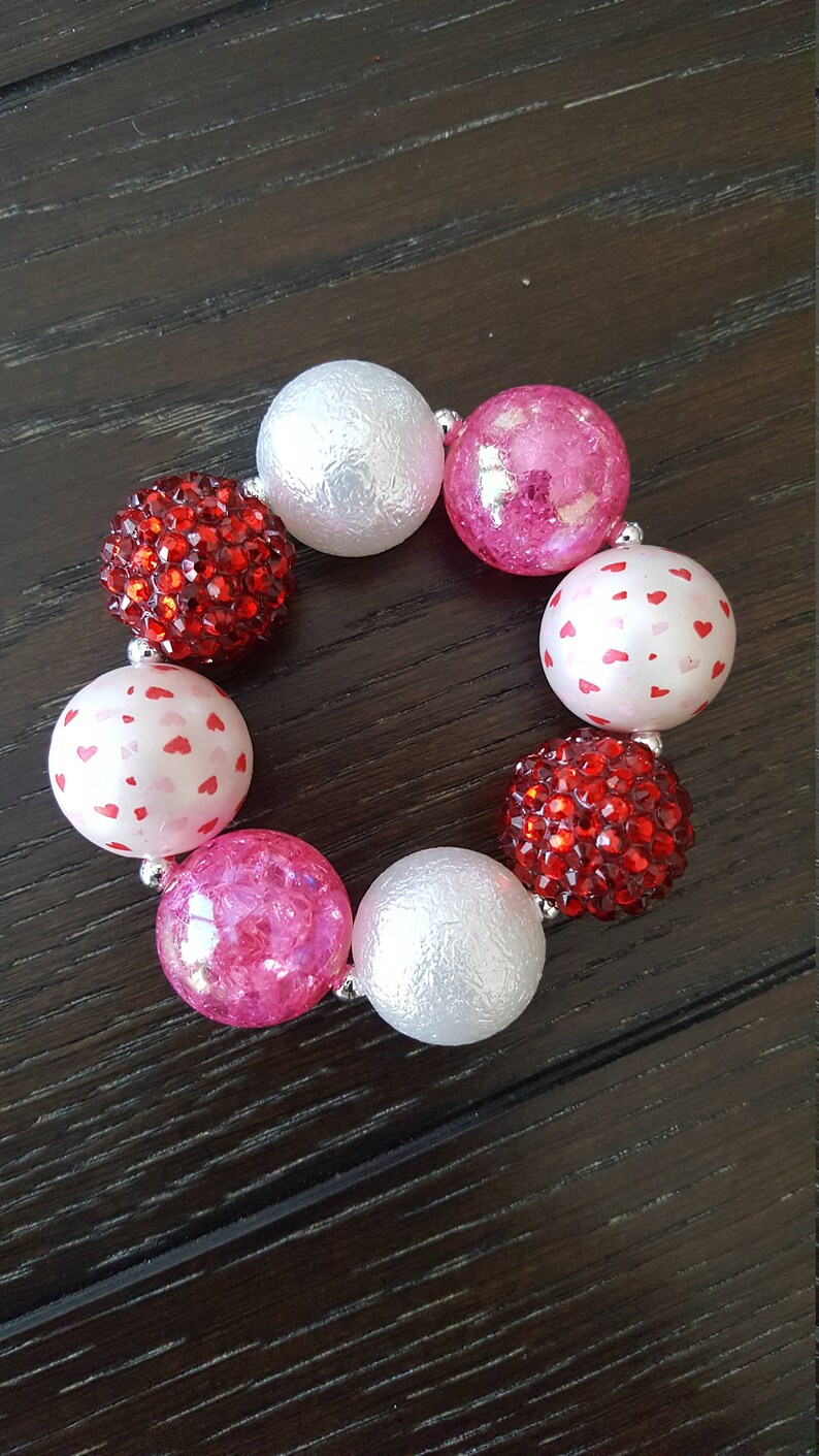 May include: A Valentine's Day themed bracelet with red and pink beads. The beads are made of different materials, including glitter, hearts, and textured plastic.